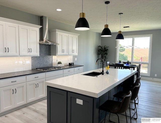 kitchen with wall chimney exhaust hood, sink, white cabinetry, hanging light fixtures, and a kitchen island with sink