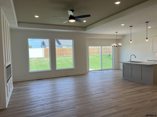 unfurnished living room with sink, light wood-type flooring, ceiling fan with notable chandelier, and a tray ceiling