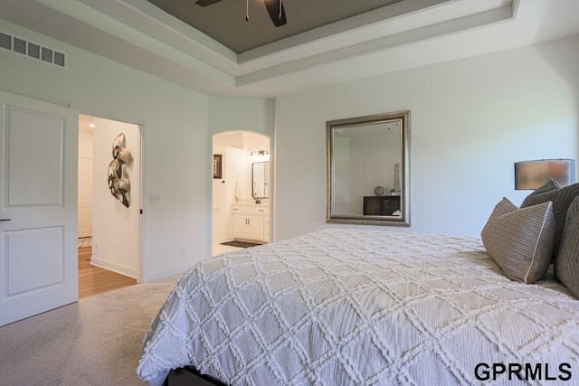 bedroom featuring connected bathroom, light carpet, ceiling fan, and a tray ceiling