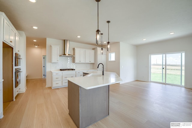 kitchen featuring white cabinetry, sink, hanging light fixtures, wall chimney range hood, and a center island with sink