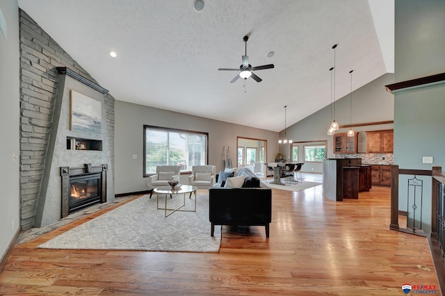 living room with high vaulted ceiling, ceiling fan with notable chandelier, a textured ceiling, a fireplace, and light hardwood / wood-style floors