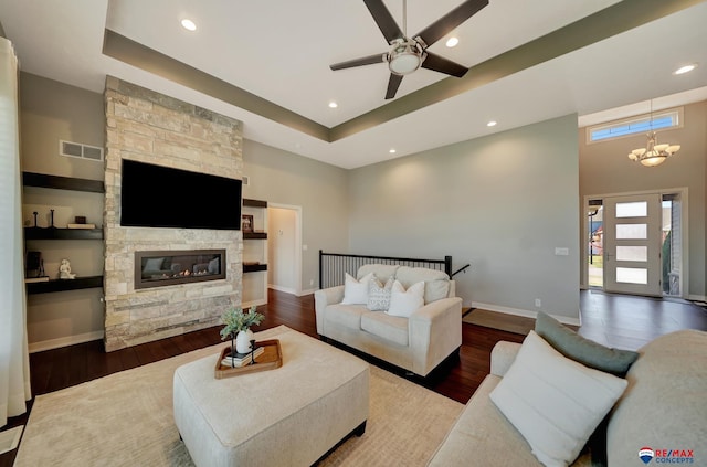 living room with a high ceiling, a raised ceiling, a stone fireplace, ceiling fan with notable chandelier, and hardwood / wood-style flooring