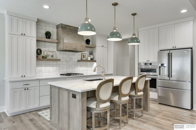 kitchen featuring premium range hood, stainless steel appliances, sink, white cabinetry, and an island with sink