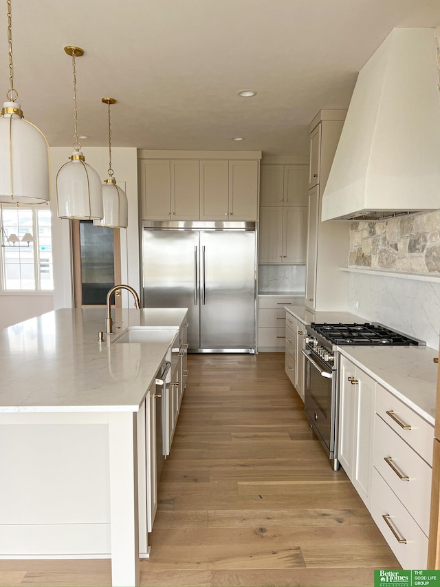 kitchen featuring custom exhaust hood, a center island with sink, hanging light fixtures, tasteful backsplash, and premium appliances