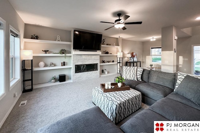 living room featuring carpet flooring, built in features, ceiling fan, and a stone fireplace