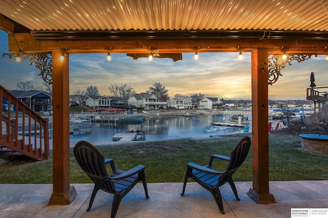 patio terrace at dusk featuring a boat dock, a water view, and a lawn