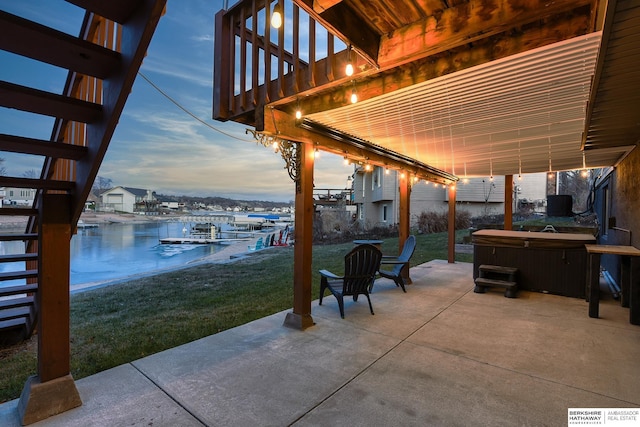 patio terrace at dusk featuring a dock, a water view, a hot tub, and a lawn