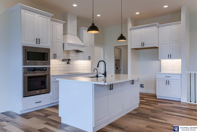 kitchen with appliances with stainless steel finishes, custom exhaust hood, a kitchen island with sink, decorative light fixtures, and white cabinetry