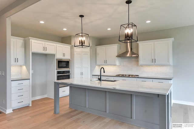 kitchen with white cabinetry, appliances with stainless steel finishes, wall chimney range hood, and an island with sink