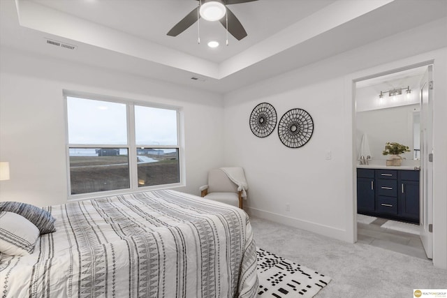 bedroom featuring a raised ceiling, light colored carpet, ensuite bath, and ceiling fan