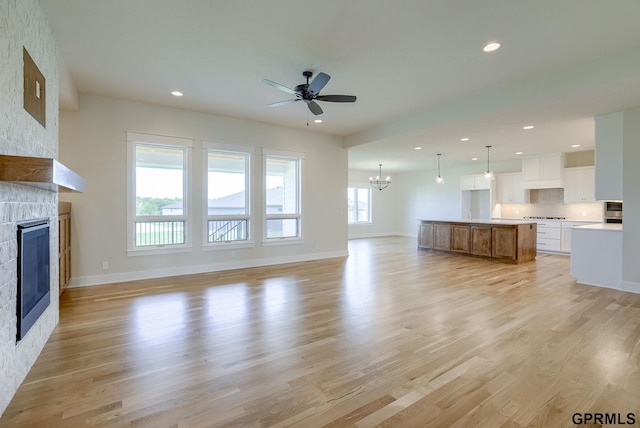 unfurnished living room with ceiling fan with notable chandelier, light hardwood / wood-style floors, a stone fireplace, and a wealth of natural light