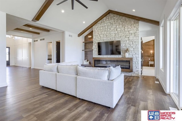 living room with ceiling fan, dark wood-type flooring, a stone fireplace, vaulted ceiling with beams, and plenty of natural light