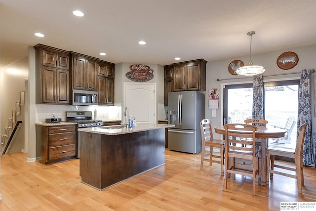 kitchen with light stone countertops, light wood-type flooring, an island with sink, dark brown cabinetry, and stainless steel appliances