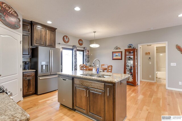 kitchen with light wood-type flooring, light stone counters, stainless steel appliances, sink, and an island with sink