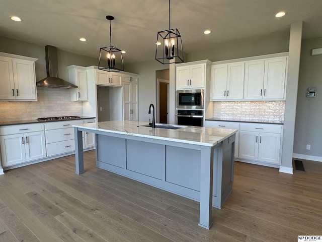 kitchen with stainless steel appliances, sink, wall chimney range hood, a center island with sink, and white cabinetry