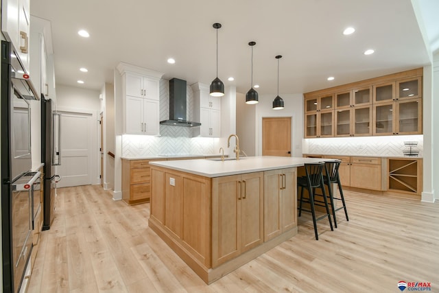 kitchen featuring sink, wall chimney range hood, decorative light fixtures, a center island with sink, and white cabinets