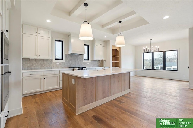 kitchen featuring decorative light fixtures, white cabinetry, premium range hood, and an island with sink