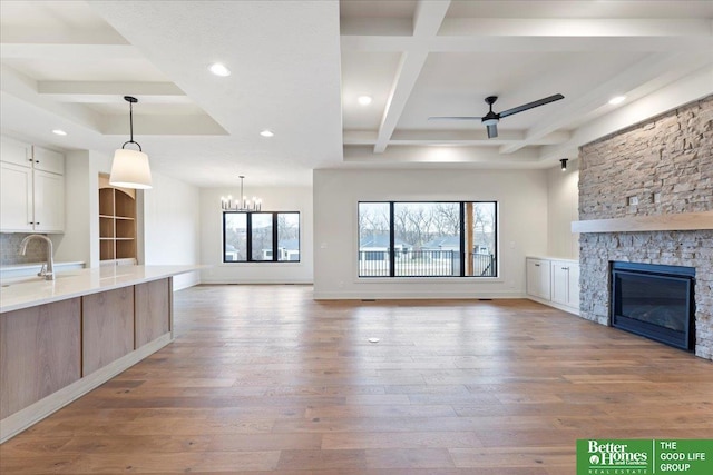 unfurnished living room with coffered ceiling, ceiling fan with notable chandelier, a stone fireplace, light hardwood / wood-style flooring, and beam ceiling