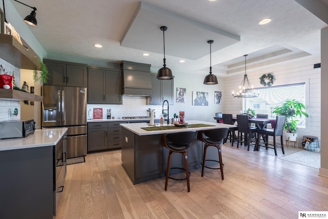 kitchen featuring premium range hood, stainless steel appliances, a tray ceiling, pendant lighting, and a center island with sink