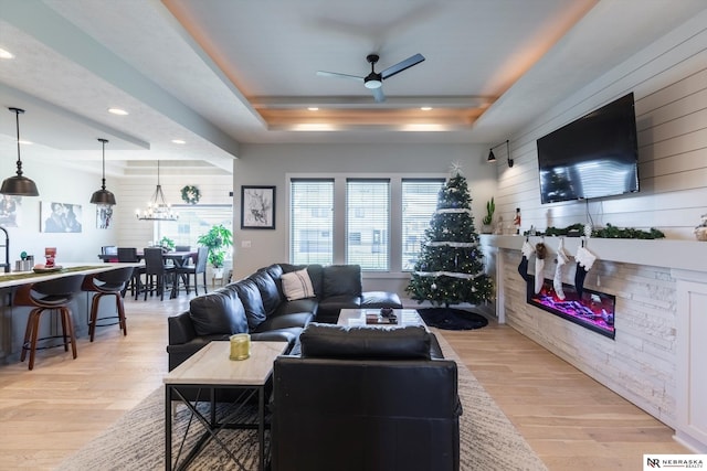 living room with ceiling fan with notable chandelier, light hardwood / wood-style floors, a raised ceiling, and wooden walls