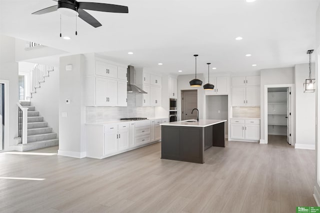 kitchen featuring a center island with sink, wall chimney exhaust hood, white cabinets, and decorative light fixtures