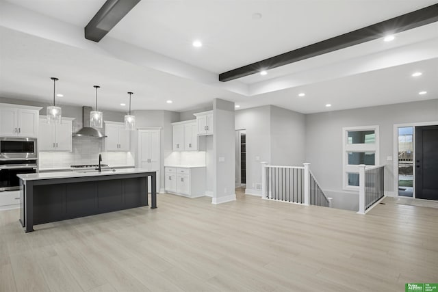 kitchen with white cabinetry, stainless steel appliances, wall chimney range hood, beamed ceiling, and an island with sink