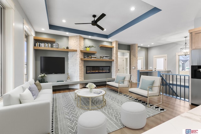 living room with a large fireplace, ceiling fan with notable chandelier, light hardwood / wood-style floors, and a tray ceiling