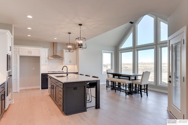 kitchen with white cabinetry, an island with sink, light wood-type flooring, pendant lighting, and wall chimney exhaust hood