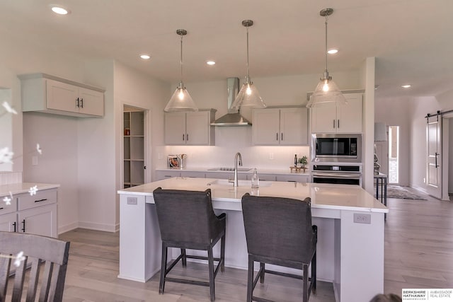 kitchen with a center island with sink, wall chimney range hood, a barn door, appliances with stainless steel finishes, and decorative light fixtures