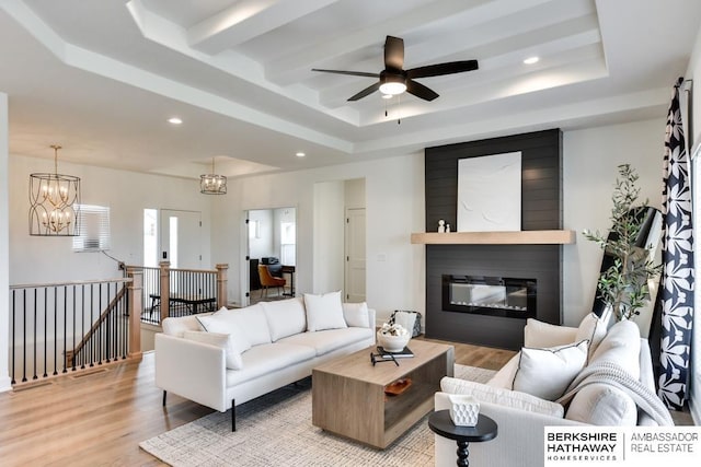 living room featuring a tray ceiling, a fireplace, ceiling fan with notable chandelier, and light wood-type flooring