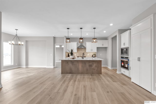 kitchen featuring wall chimney exhaust hood, stainless steel oven, built in microwave, a kitchen island with sink, and white cabinetry