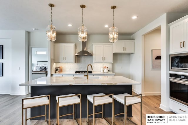 kitchen featuring pendant lighting, a center island with sink, wall chimney exhaust hood, white cabinetry, and stainless steel appliances