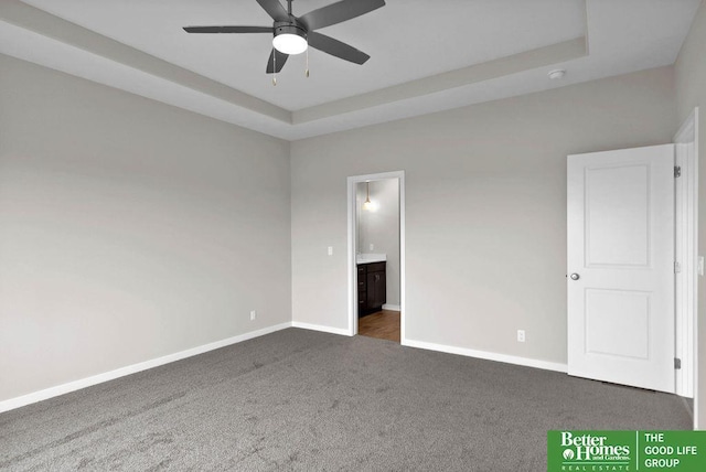 unfurnished bedroom featuring a tray ceiling, connected bathroom, ceiling fan, and dark carpet