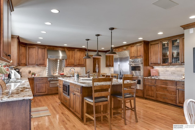 kitchen featuring hanging light fixtures, wall chimney range hood, built in appliances, a center island with sink, and light wood-type flooring