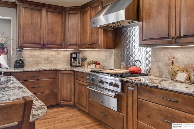 kitchen with wall chimney exhaust hood, decorative backsplash, light stone countertops, light wood-type flooring, and stainless steel gas cooktop