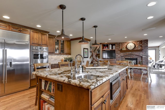 kitchen with a brick fireplace, a kitchen island with sink, sink, built in appliances, and decorative light fixtures