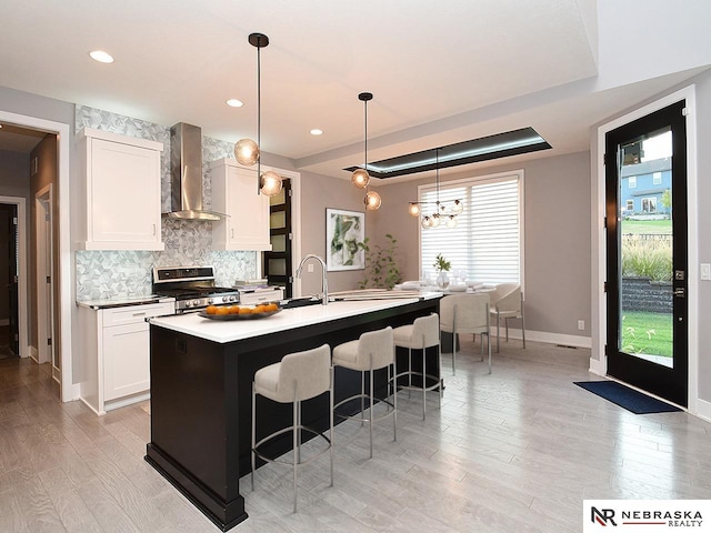 kitchen featuring hanging light fixtures, stainless steel gas range, wall chimney exhaust hood, an island with sink, and white cabinetry