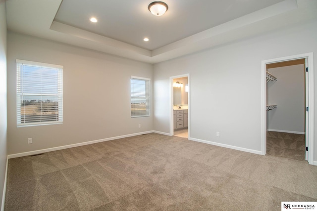 unfurnished bedroom featuring a tray ceiling, ensuite bath, light colored carpet, and a spacious closet