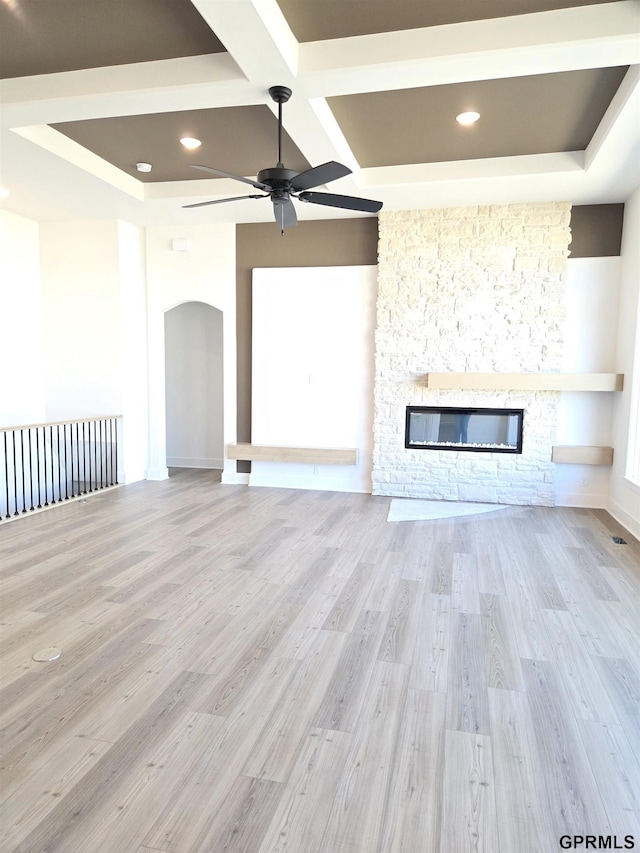 unfurnished living room with wood finished floors, coffered ceiling, arched walkways, ceiling fan, and a stone fireplace