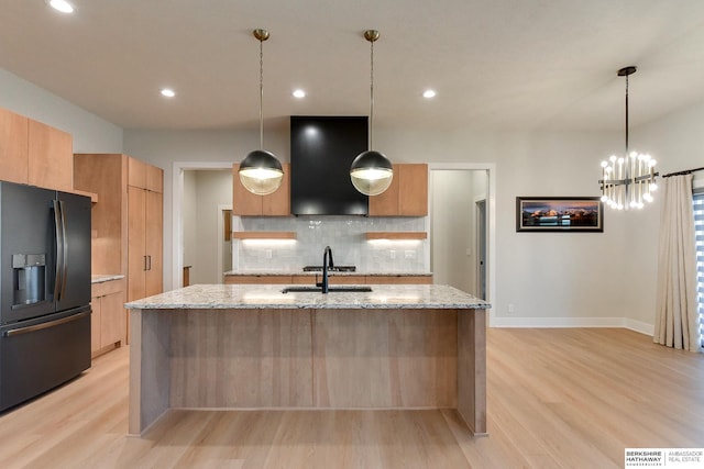 kitchen with a kitchen island with sink, black refrigerator with ice dispenser, ventilation hood, decorative light fixtures, and light stone counters