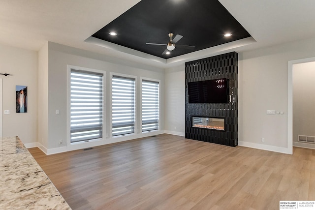 unfurnished living room featuring ceiling fan, a large fireplace, light wood-type flooring, and a tray ceiling