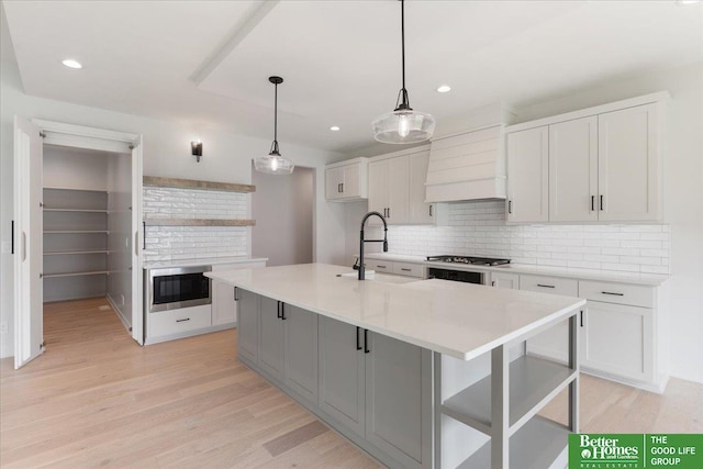 kitchen with light wood-type flooring, tasteful backsplash, custom range hood, a kitchen island with sink, and white cabinetry