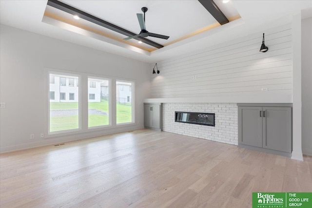 unfurnished living room with a brick fireplace, ceiling fan, light wood-type flooring, and a tray ceiling