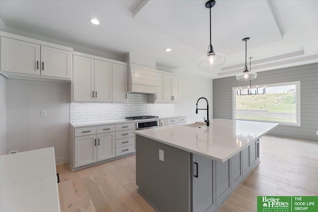 kitchen with sink, light stone counters, a raised ceiling, an island with sink, and hanging light fixtures