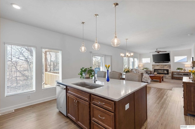 kitchen with dishwasher, a center island with sink, sink, a stone fireplace, and ceiling fan with notable chandelier