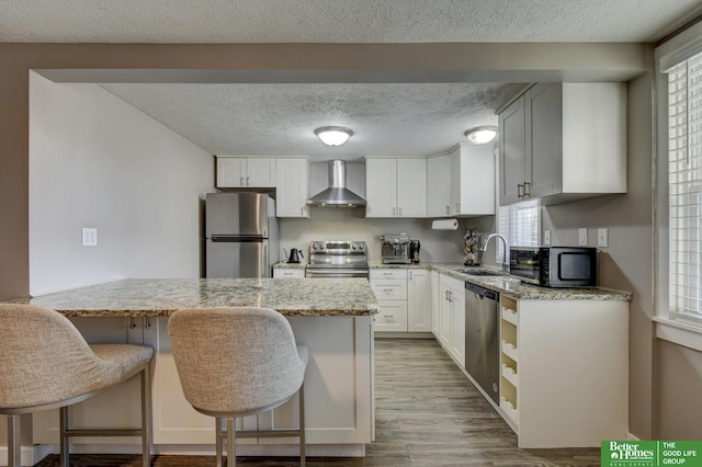 kitchen featuring a kitchen bar, stainless steel appliances, white cabinets, wall chimney exhaust hood, and light stone counters