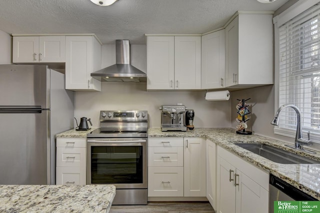 kitchen with light stone countertops, appliances with stainless steel finishes, white cabinetry, wall chimney range hood, and sink