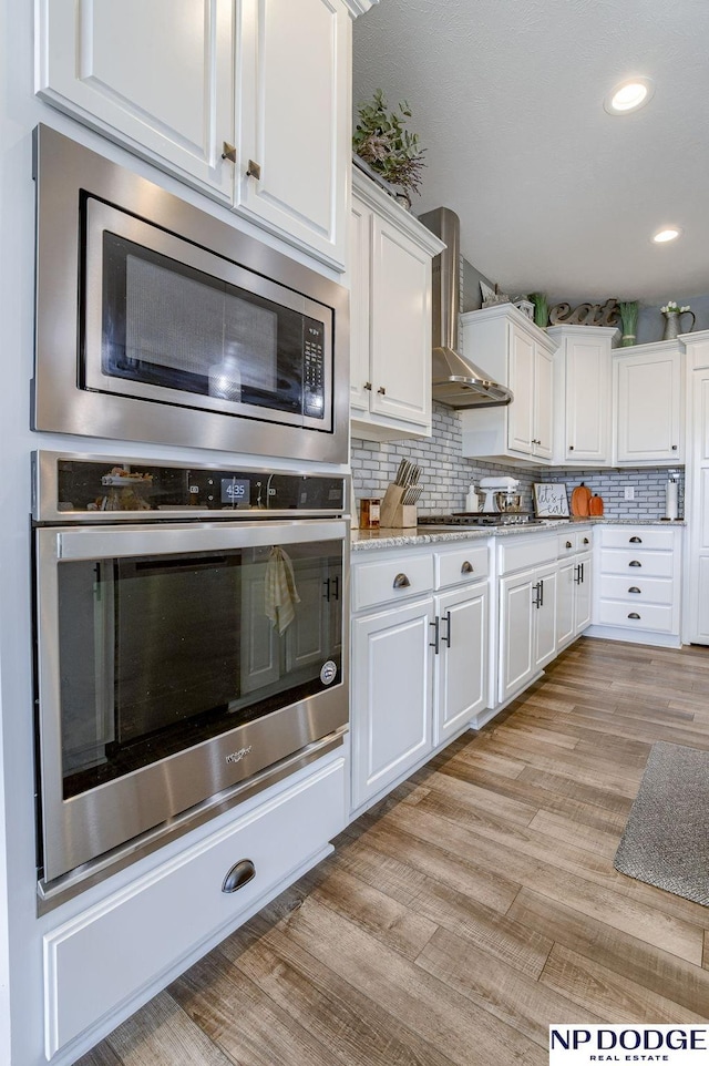 kitchen featuring backsplash, light hardwood / wood-style floors, white cabinetry, appliances with stainless steel finishes, and wall chimney exhaust hood