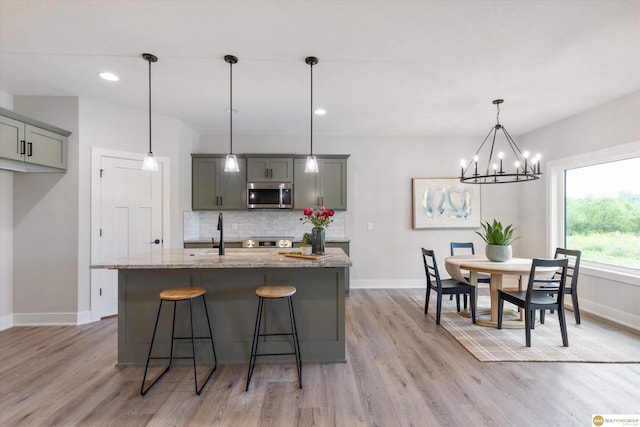 kitchen featuring a center island with sink, backsplash, decorative light fixtures, light wood-type flooring, and light stone countertops