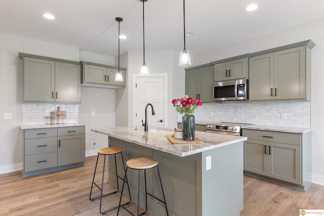 kitchen with appliances with stainless steel finishes, decorative backsplash, a kitchen island with sink, hanging light fixtures, and light stone counters